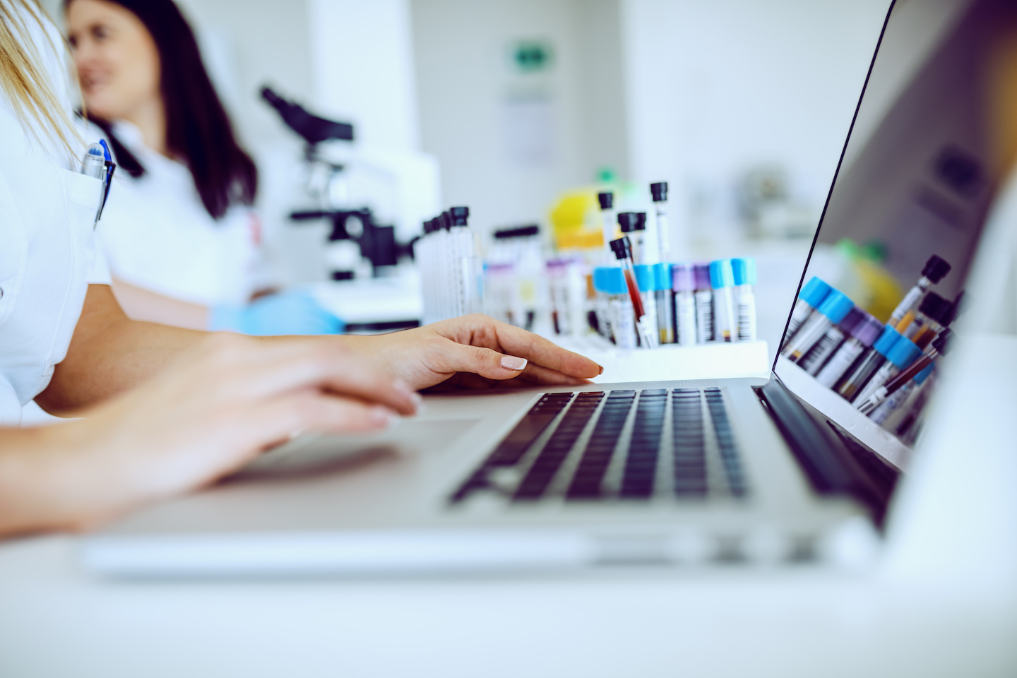 Close up of female lab assistant in white uniform sitting in lab and using laptop for data entry. In background is her colleague working. Entering sample information into laptop